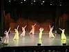 Sju dansere i lyse kostymer opptrer på en scene med mørk bakgrunn og scenelys.