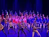 Stor gruppe dansere opptrer på en opplyst scene med blå og lilla bakgrunn.