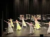Flere dansere i lyse kostymer opptrer på en mørk scenen foran et svart sceneteppe.