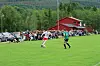 Fotballspillere på en gressbane med publikum langs sidelinjen og en rød bygning bak.