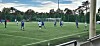 Fotballspillere i blå og hvite drakter spiller på en kunstgressbane ved et mål.