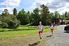 To løpere på en grusvei ved en grønn park med trær, en rød bygning og en bil i bakgrunnen.