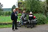 Tre personer står ved en motorsykkel med sidevogn på en grusvei foran trær og gress.