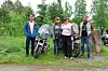 Fire personer står samlet ved en motorsykkel på en grusplass omgitt av trær og vegetasjon.