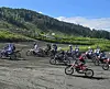 Flere motorsykler kjører på en grusplass foran en grønn ås under klar himmel.