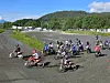 Motocrossførere kjører gjennom en sving på en grusbane med grønn skog og åser bak.