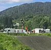 Syklist i luften over en jordvoll med skogkledd ås og folk og vogner i bakgrunnen.