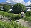 To motocrossførere kjører på en bane gjennom grønn vegetasjon med hus og fjell i bakgrunnen.