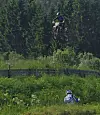 Motorsyklist hopper høyt over en grønn bakke med skog i bakgrunnen.