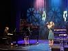 Sanger og pianist på en scene med flygel, mikrofoner og blå belysning.