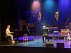 Pianist på scene med blå og lilla scenelys, flygel, trommer og annet musikkutstyr i bakgrunnen.