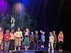 Barn står oppstilt på en scene med fargede scenelys og mørk bakgrunn.