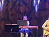 Musiker ved keyboard på scene med mørk lyssetting og abstrakt bakgrunn.