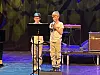 To personer står på en scene med mikrofon, notestativ og musikkinstrumenter i bakgrunnen.