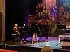 Pianist ved et svart flygel på en scene med mikrofonstativ og stol i varmt og blått lys.