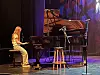 Pianist sitter ved et flygel på en opplyst scene med mikrofonstativ og krakk.
