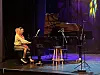 To personer spiller piano sammen ved et flygel på en scene med lilla lys.