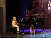 En pianist sitter ved et svart flygel på en scenen med varme lys og mikrofonstativ.