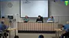 Tre personer ved et panelbord i et møterom med publikum og prosjektorlerret i bakgrunnen.