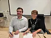 To personer sitter ved et bord med dokumenter og penner foran en projektorskjerm.