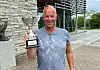 Mann i blå T-skjorte holder en pokal foran en bygning med steinvegg.