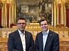 To menn i dress står foran et stort maleri og en gullfarget dekorativ bakgrunn.