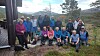 En gruppe personer står og sitter samlet utendørs i et fjellandskap med trær og utsikt i bakgrunnen.