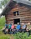 Seks turkledde personer sitter på en benk ved en gammel tømmerløe i skogen og spiser matpause.