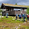 Flere turgåere med ryggsekker går opp en bratt bakke mot en hytte.