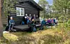 En gruppe turkledde personer ved en tømmerhytte omgitt av trær og gresskledd terreng.