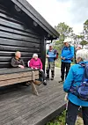 Personer i turklær sitter og står ved en trehytte på en platting i skogsterreng.