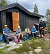 Fem turkleddee personer sitter og hviler ved en trehytte med tursekker og flasker foran seg.