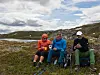 Tre personer sitter på stoler i lynghei ved et vann under en skyet himmel.