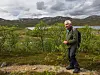 En mann står på en sti ved lav vegetasjon, med fjell, vann og overskyet himmel i bakgrunnen.