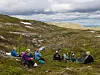 Flere turgåere sitter i en skråning med lyng, stein og små trær i et fjellområde.