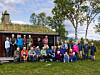 Stor gruppe mennesker samlet foran en hytte på en gressplen med trær i bakgrunnen.
