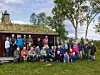 Stor gruppe mennesker samlet foran en hytte på en gressplen med trær i bakgrunnen.