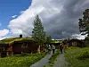 To personer går på grussti mellom trehytter under mørke skyer.