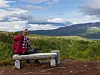 Person med rød ryggsekk sitter på en benk og ser ut over grønn natur og fjell.