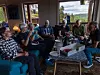 Flere eldre personer sitter i sofaer rundt et bord i en stue med store vinduer og fjellutsikt.