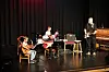 Tre musikere spiller på en scene med trommesett, gitarer og piano i et mørkt lokale.