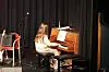 Person spiller piano på en liten scene med mørk bakgrunn og mikrofonstativ.
