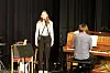 En sanger ved mikrofon og en pianist ved piano på en mørk scene.