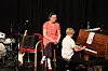 To personer ved et piano på en scene, med trommesett og gitar i bakgrunnen.