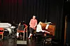 Musikere på en mørk scene med piano, trommesett og lydutstyr under øving.