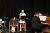 Sanger på scene ved mikrofon, med pianist ved piano og musikkinstrumenter i bakgrunnen.