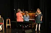 To unge jenter og en dame på en scene, én synger ved et piano mens to andre sitter ved instrumentet.