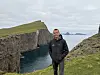 Mann står på gresskledd klippekant med bratte sjøklipper og hav i bakgrunnen.