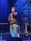 Artist synger på scene med mikrofon, gitar og trommesett i blå belysning.