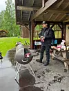 En mann står ved en liten grill under et tak av tre i en park, omgitt av bord med mat og utstyr.